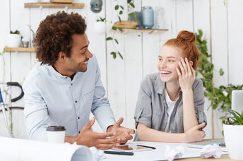 Beautiful young redhead European woman designer with hair bun laughing at jokes of her cheerful charismatic dark-skinned male colleague
