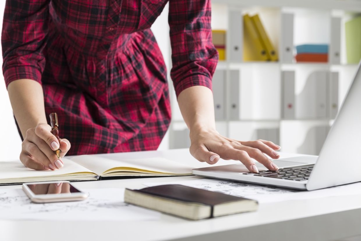 Close up of woman in checkered dress typing on her laptop while standing and making notes Close up of woman in checkered dress typing on her laptop while standing and making notes