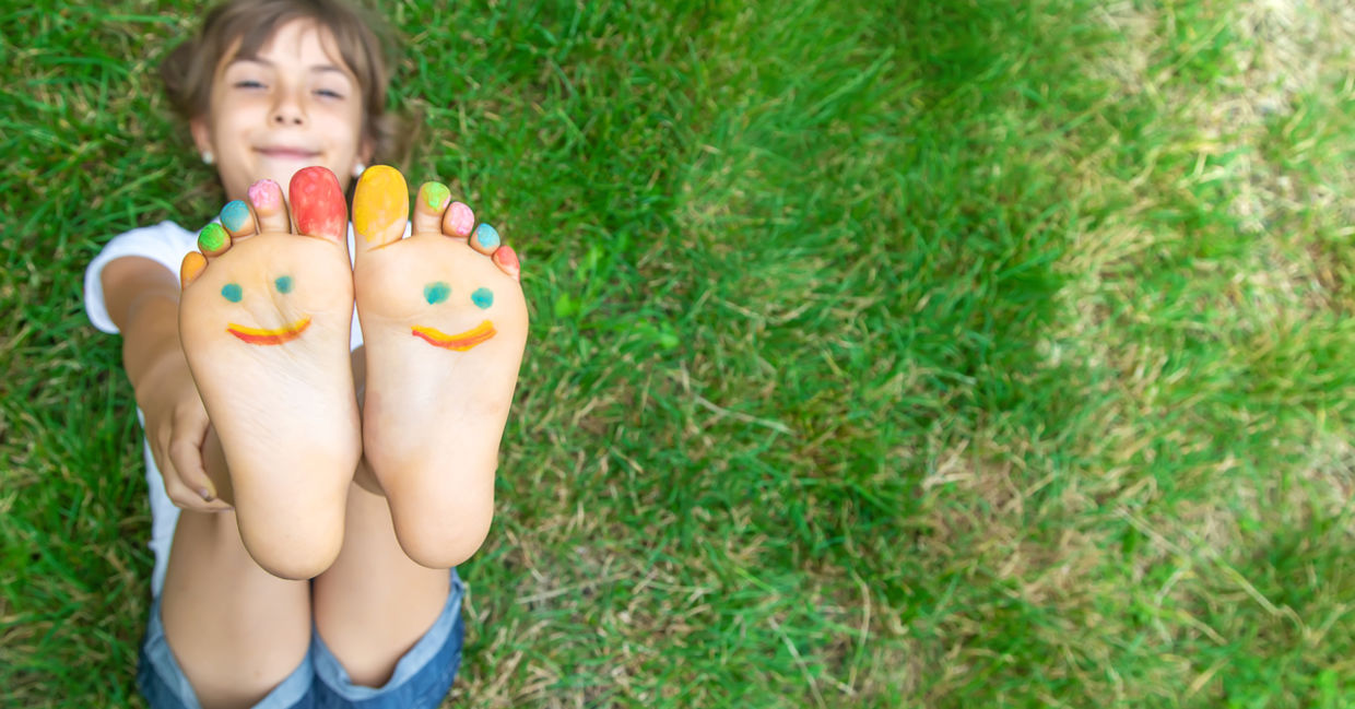A child lies on the grass, feet up, with painted toes and a happy face on her feet. A child lies on the grass, feet up, with painted toes and a happy face on her feet.