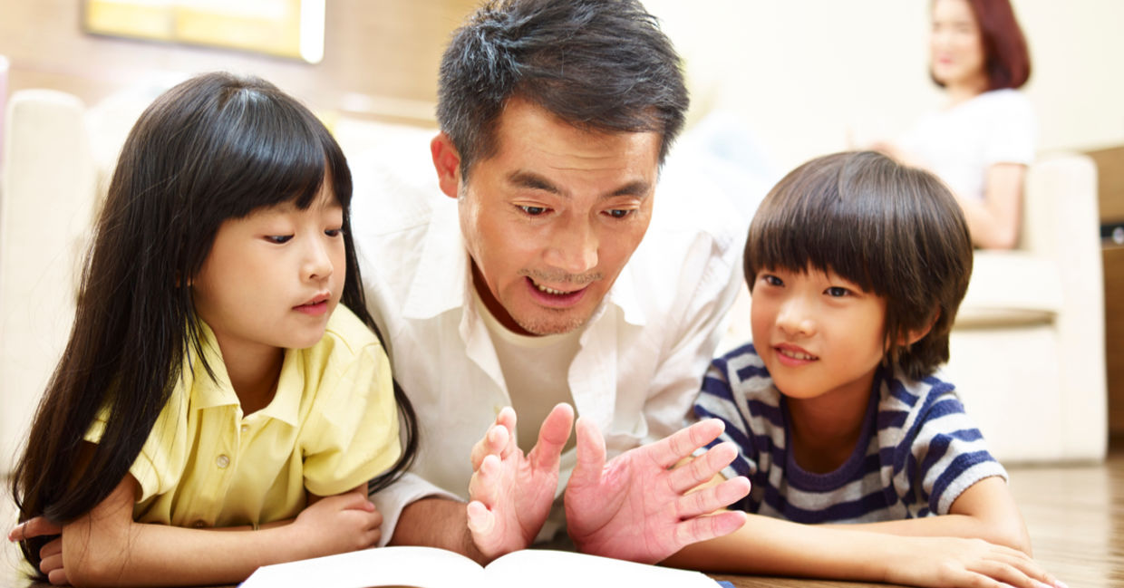 A father lies on the floor reading a book to his two children. A father lies on the floor reading a book to his two children.