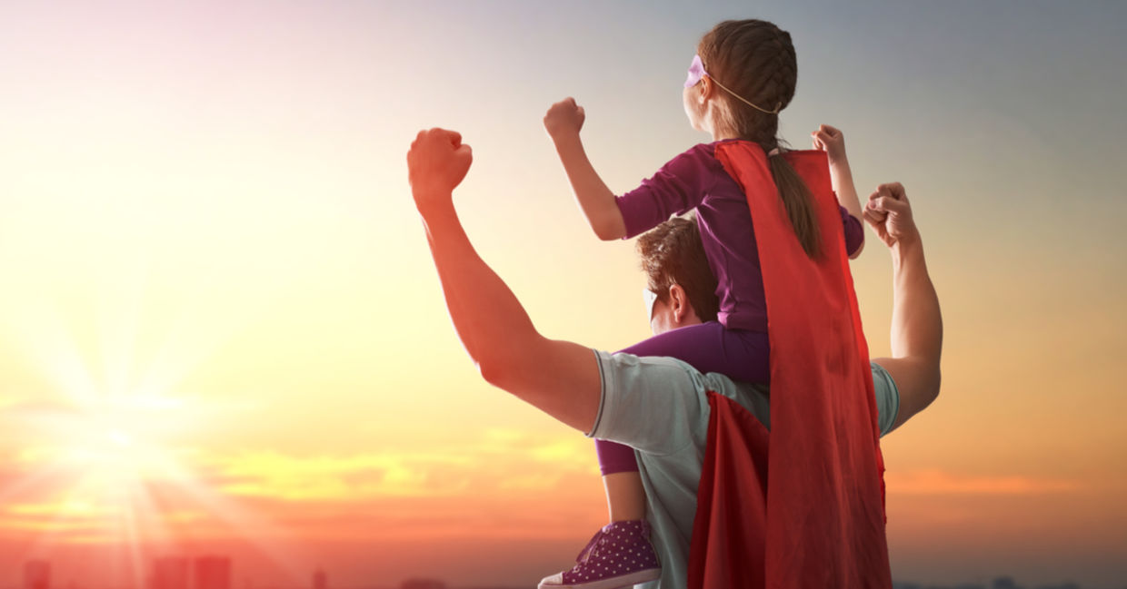 A father holds his daughter on his shoulders, both of them wearing empowering superhero capes. A father holds his daughter on his shoulders, both of them wearing empowering superhero capes.