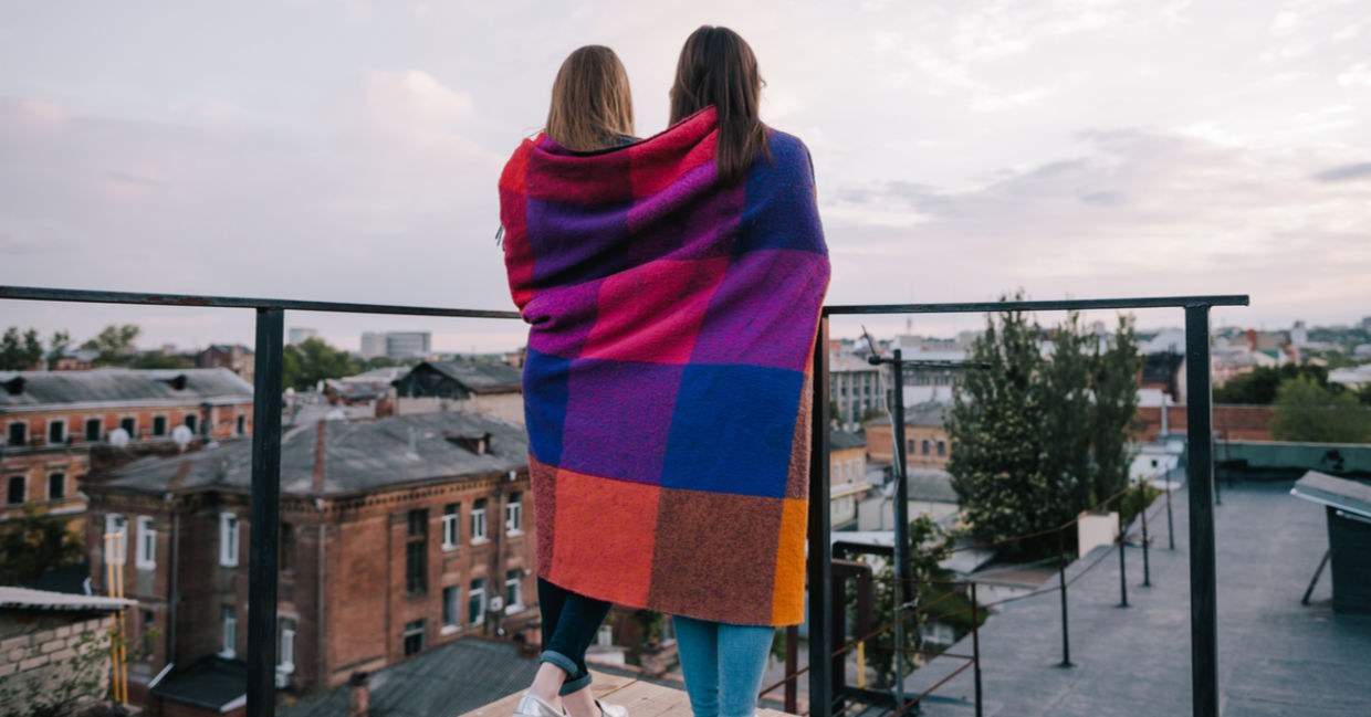 Two friends wrapped in a blanket stand outside on a balcony. Two friends wrapped in a blanket stand outside on a balcony.