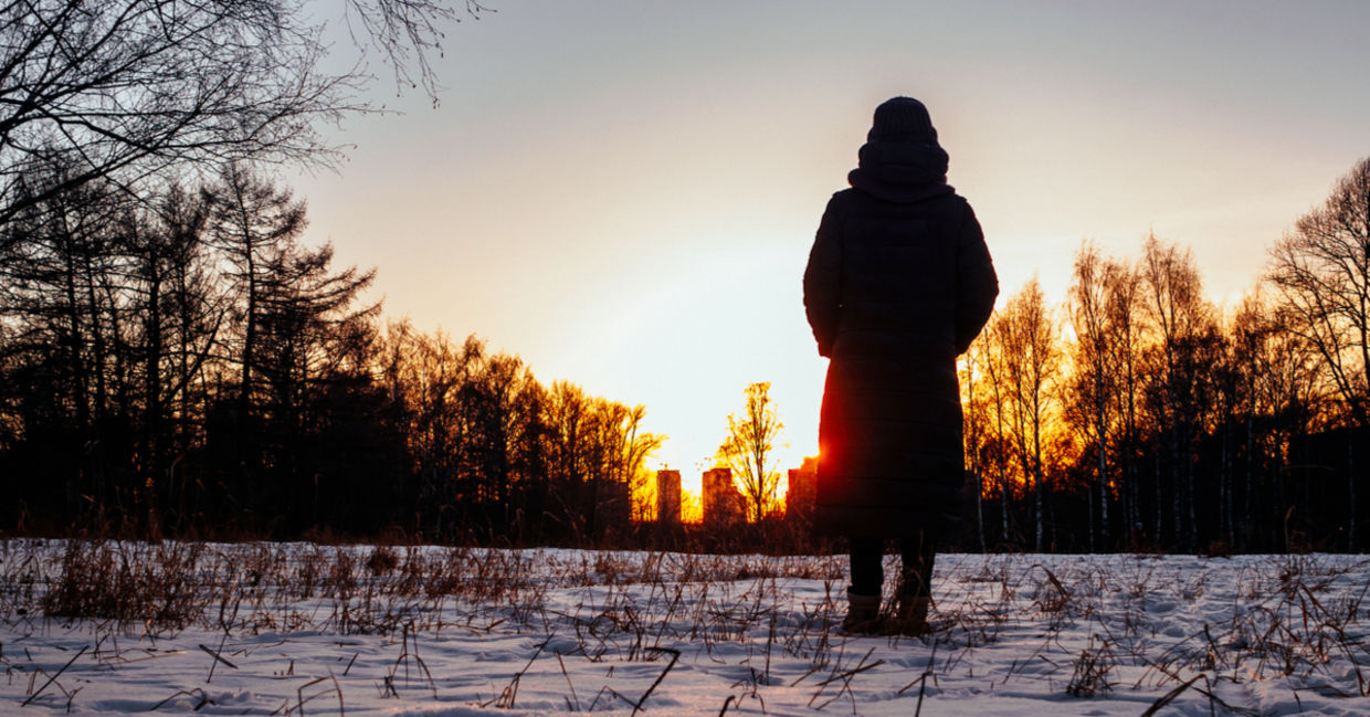 A woman stands among trees in the snow watching the sun setting behind buildings. A woman stands among trees in the snow watching the sun setting behind buildings.